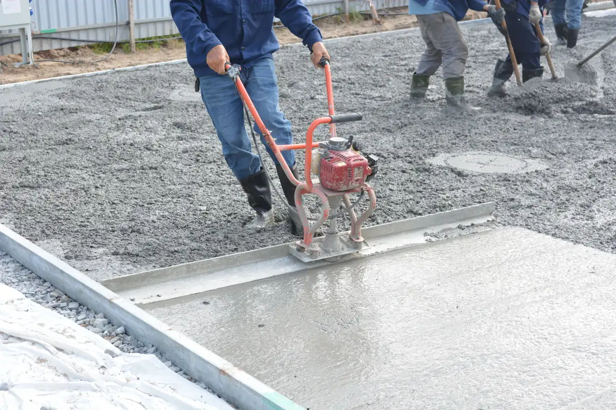 Finisher running a power screed across a fresh concrete slab between steel forms while the crew spreads concrete behind
